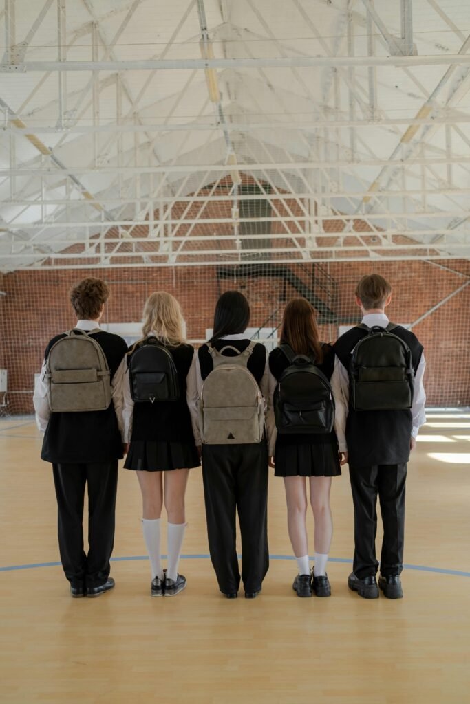 A group of five students in uniform stand indoors with backpacks, facing away.